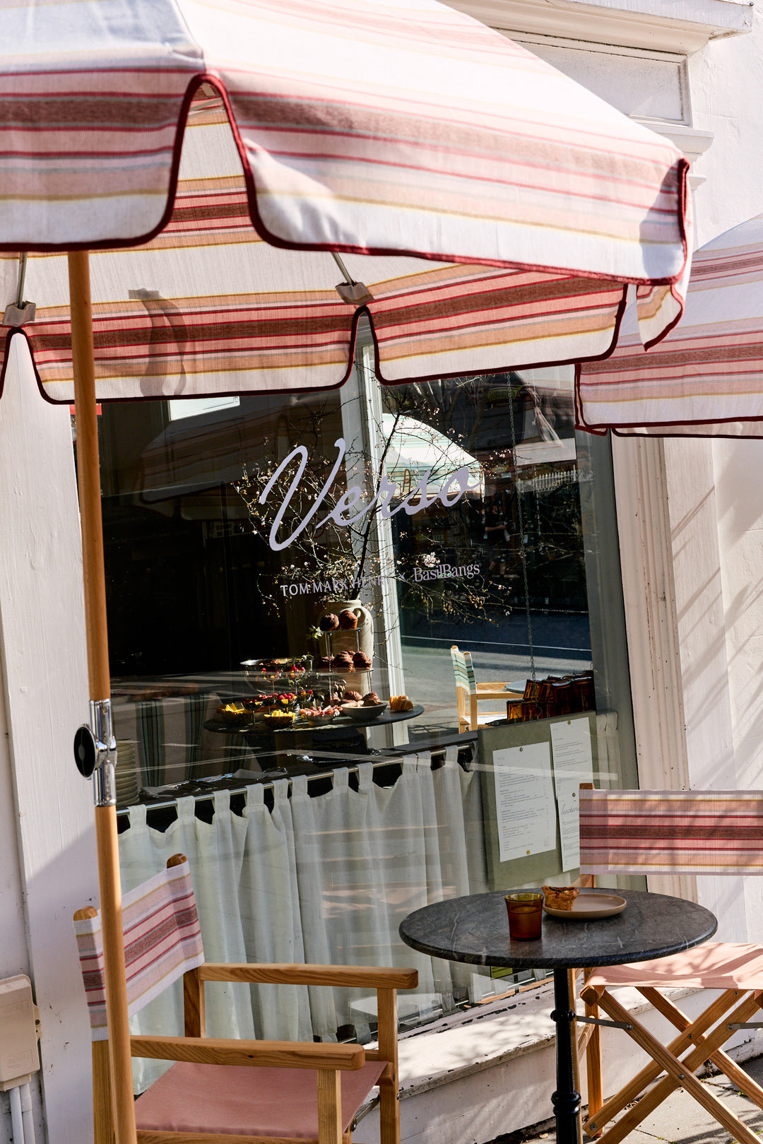A cafe patio scene with striped umbrellas, wooden chairs, a small round table with drinks, and a glass window reflecting outdoor light. The window has the word 'Venus' in white script.