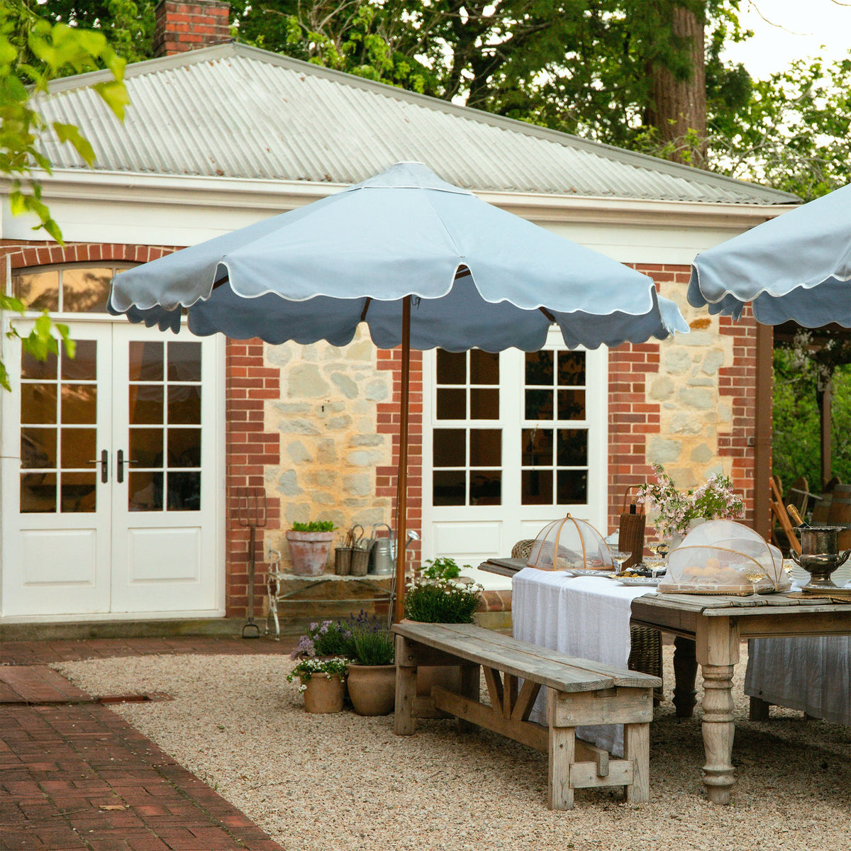 Outdoor patio with large blue patio umbrellas, wooden table, and bench in front of a heritage brick house.
