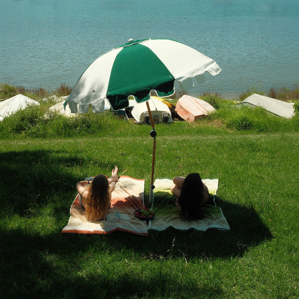 A forest green and white Basil Bangs Luxury Parasol shades two people relaxing on thick cotton beach towels on a grassy hill overlooking a calm bay.
