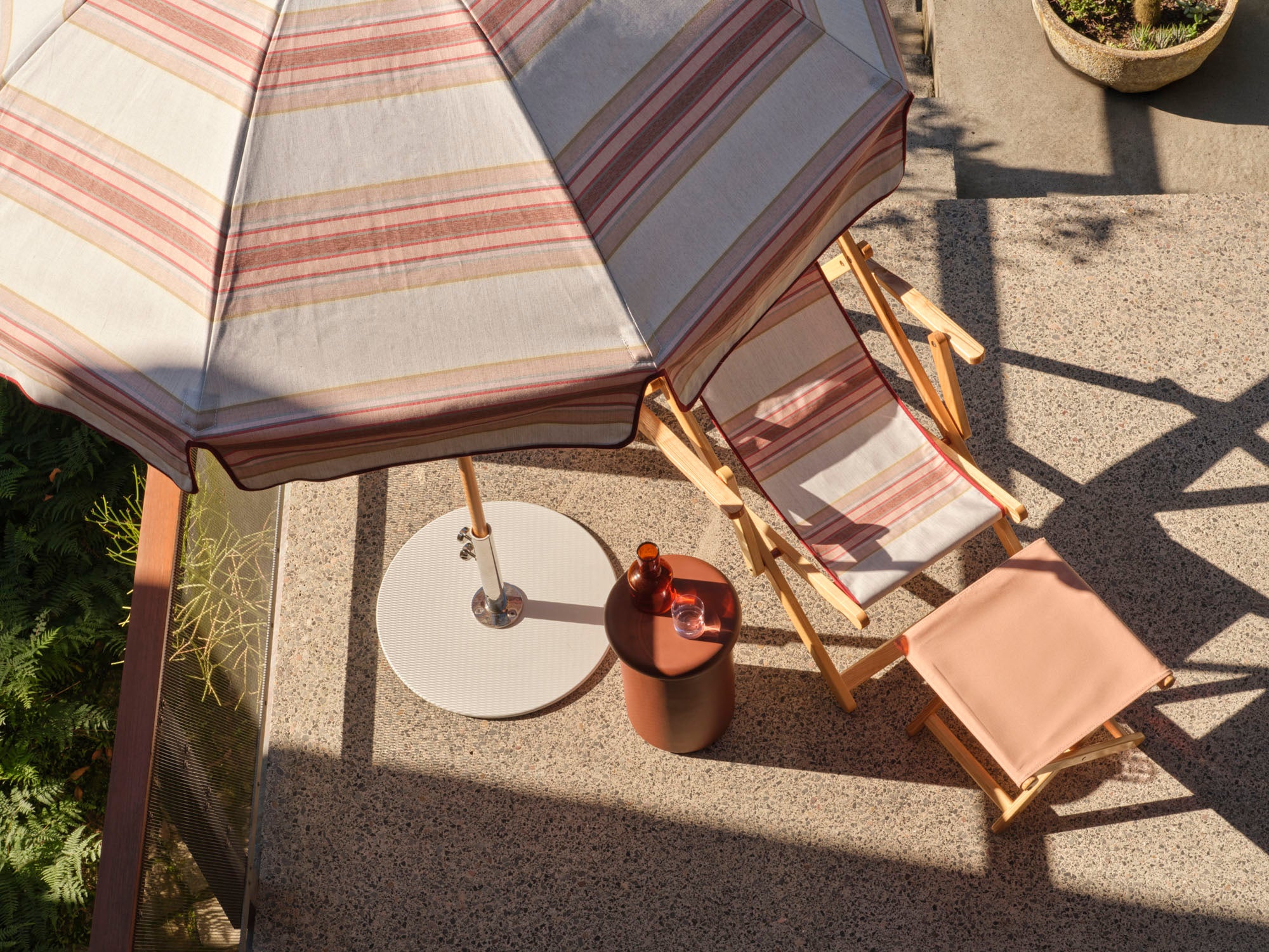 A styled outdoor patio scene featuring a striped beach umbrella, a wooden folding lounge chair, a matching ottoman, and a small round side table on a textured concrete patio.
