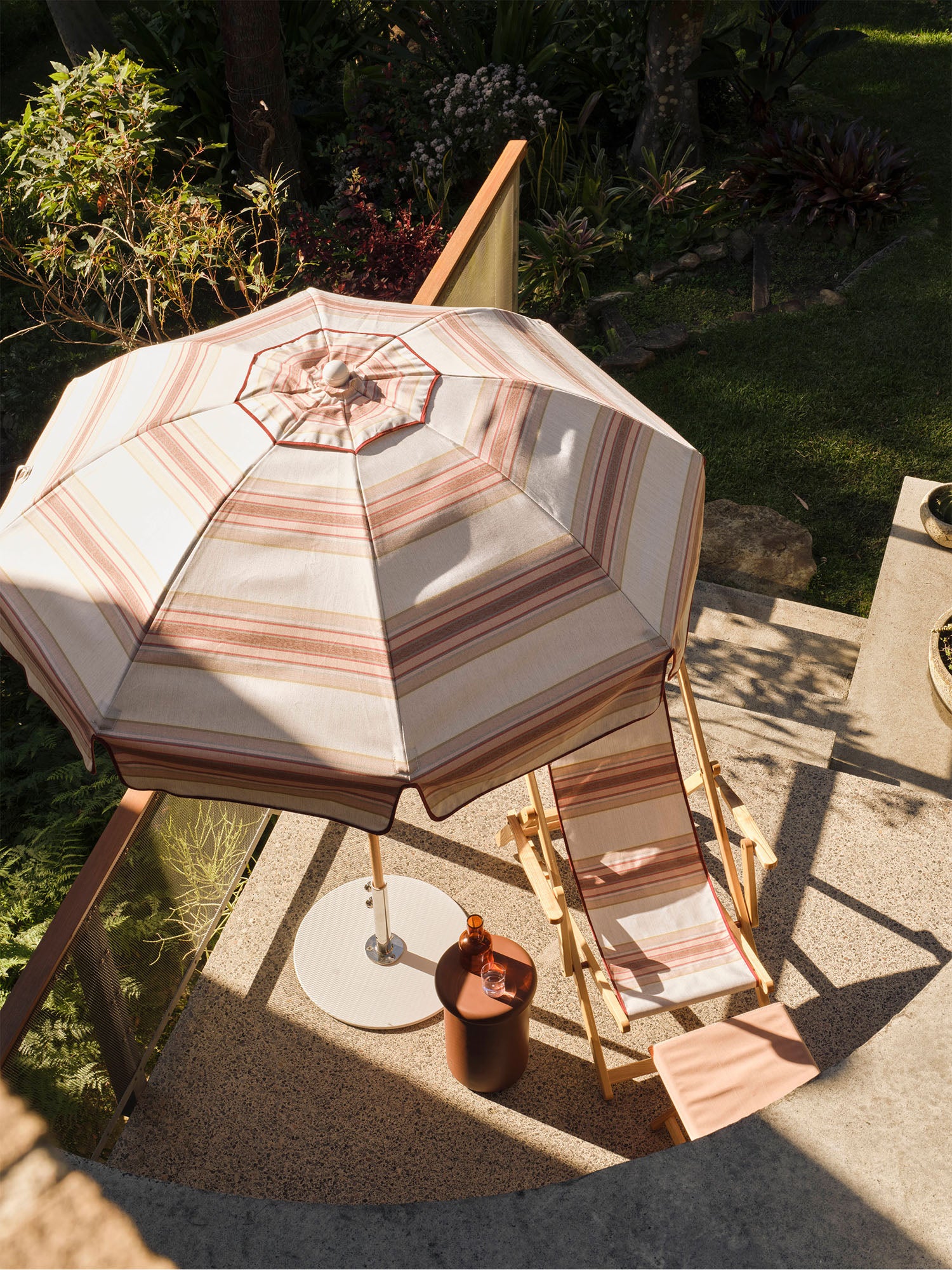 A striped patio umbrella in beige, pink, and brown tones is set up on a stone patio next to a matching timber lounge chair and a small side table with a bottle, with a garden background.